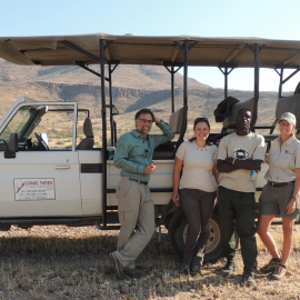 A rhino ranger is being a guide for some tourists, they are standing outside their vehicle in the desert landscape